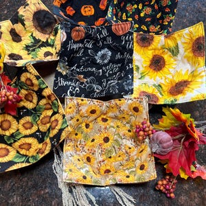 May include: A collection of six fabric bowl cozies featuring various seasonal patterns, including orange pumpkins on black, sunflowers on yellow, and black fabric with white text reading Fall, Autumn, and thankful, arranged in a circular display.