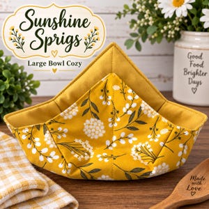 May include: A large bowl cozy featuring a mustard yellow fabric with a white floral sprig pattern and a solid mustard yellow interior. The item is labeled Sunshine Sprigs Large Bowl Cozy with a decorative graphic and text on the fabric.