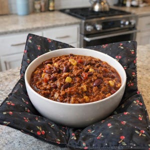 May include: A white ceramic bowl filled with chili containing ground meat, red kidney beans, and yellow corn kernels. The bowl sits inside a black quilted fabric bowl cozy featuring a repeating pattern of small red cherries and green leaves.