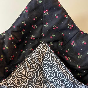 May include: A quilted fabric bowl cozy featuring a black interior with a red cherry and white dot pattern, and an exterior with a black and white swirling spiral design. The bowl cozy has four distinct corners and visible stitched seams.