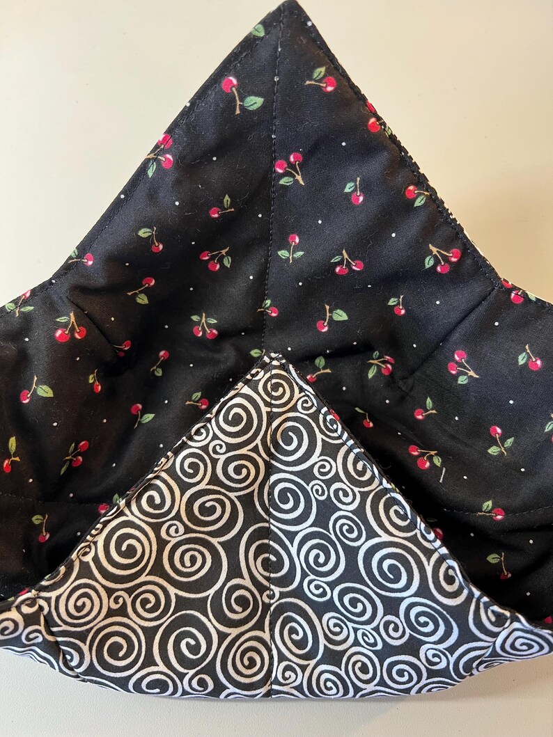 May include: A quilted fabric bowl cozy featuring a black interior with a red cherry and white dot pattern, and an exterior with a black and white swirling spiral design. The bowl cozy has four distinct corners and visible stitched seams.