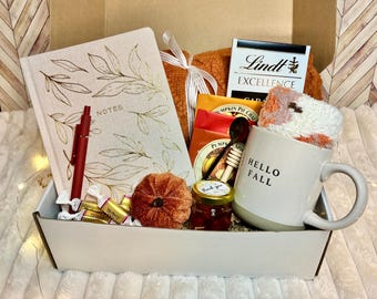 May include: A seasonal gift box containing a journal with gold leaf detailing, a red pen, a mug with "HELLO FALL", a jar of jam, chocolates, tea, a plush pumpkin, and a blanket. A perfect autumn gift.