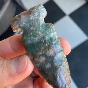 May include: A hand-held, green and brown arrowhead with a notched design. The stone artifact has a pointed tip and a textured surface, showcasing natural variations in color and pattern. The arrowhead is a relic of ancient craftsmanship.