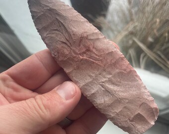 May include: A hand-held, reddish-brown arrowhead with a textured surface. The arrowhead is elongated with a pointed tip and a wider base. The stone tool appears to be hand-crafted, with visible ridges and a slightly uneven shape.
