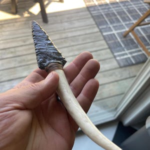 May include: A dark gray obsidian arrowhead with a white bone handle. The arrowhead is sharp and pointed, and the handle is smooth and polished.