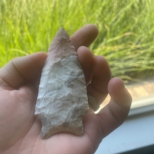 May include: A white and pink arrowhead made of stone. The arrowhead is pointed and has a rough texture.