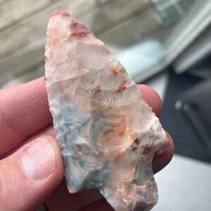 May include: A hand-held, multi-colored arrowhead. The arrowhead is primarily off-white with patches of red, orange, and blue. The stone has a rough, textured surface and a sharp point.