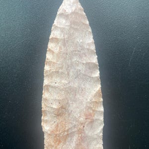 May include: A pale pink and cream-colored stone arrowhead featuring a sharp, pointed tip and serrated edges. The surface displays a textured, flaked appearance with visible mineral variations and a flat, straight base, showcasing a crafted design.