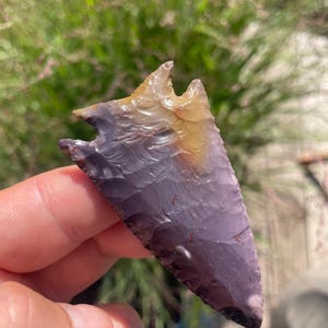 May include: A triangular stone arrowhead featuring a notched base and a pointed tip. The surface displays a smooth, flaked texture with a color gradient transitioning from deep purple at the bottom to a translucent golden yellow at the top.