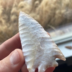 May include: A cream-colored, ancient arrowhead with a textured surface and a notched base. The arrowhead is held in a hand, showcasing its intricate craftsmanship and historical significance. The stone tool is a relic of a bygone era.