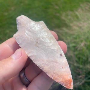 May include: A pale, translucent stone projectile point with a pointed tip and a notched base. The surface features a textured, flaked appearance with subtle white, cream, and light orange color variations throughout the crystalline structure.
