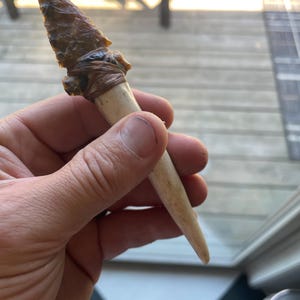 May include: A brown and tan arrowhead attached to a long, white bone handle. The arrowhead is wrapped with a brown rubber band.