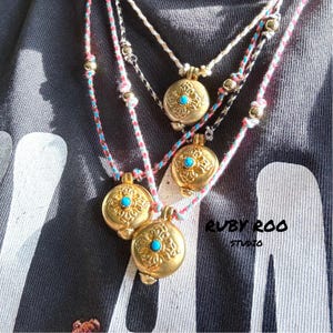 Peut inclure: Trois colliers en or avec des pierres turquoise et des motifs complexes sont suspendus à un tissu rayé noir et blanc. Les colliers sont en cordon et ont un petit pendentif rond avec une pierre turquoise au centre. Le texte "RUBY ROO STUDIO" est visible dans le coin inférieur droit de l'image.