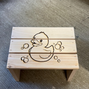May include: A light-colored wooden step stool featuring a dark, burned-in line art illustration of a rubber duck surrounded by several circular bubbles. The stool is constructed from three horizontal wooden planks with visible grain.