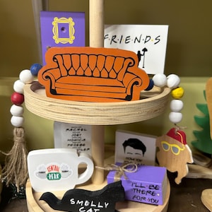 May include: A two-tiered wooden tray with decorative Friends-themed items. Includes an orange couch, a Central Perk mug, a black cat that says "Smelly Cat", and a purple box that says "I'll be there for you".