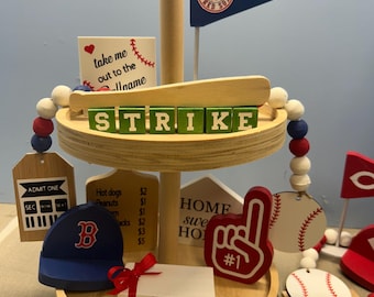 Pode incluir: Uma bandeja de madeira de dois níveis com decorações com tema de beisebol. Inclui um boné de beisebol do Boston Red Sox, um dedo de espuma, uma bola de beisebol e uma flâmula. A bandeja tem a palavra "STRIKE" escrita em blocos verdes.