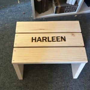 May include: A light-colored wooden step stool featuring three horizontal planks forming the top surface. The center plank is engraved with the name HARLEEN in dark, bold, capital letters. The stool has two vertical side supports.