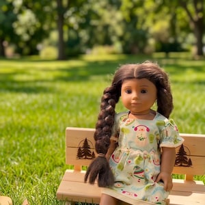 May include: A doll with long brown hair in a braid wears a light-colored dress with a colorful pattern of small icons. The doll sits on a light-colored wooden bench featuring two small pine tree engravings, next to a small wooden campfire cutout.