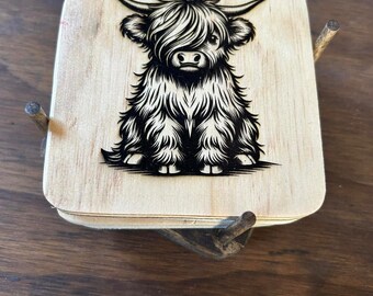 Puede incluir: Un juego de cuatro posavasos de madera con una ilustración en blanco y negro de una vaca de las tierras altas. Los posavasos están apilados uno encima del otro y tienen un aspecto rústico y hecho a mano.