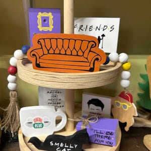 May include: A two-tiered wooden tray with decorative Friends-themed items. Includes an orange couch, a Central Perk mug, a black cat that says "Smelly Cat", and a purple box that says "I'll be there for you".