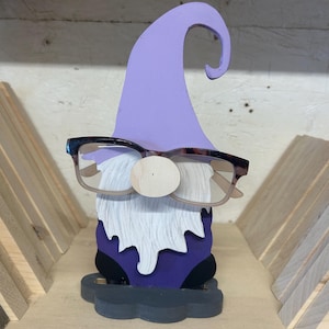 May include: A wooden gnome figurine with a lavender hat and a purple body. The gnome has a white beard, a wooden nose, and wears eyeglasses. The figurine is on a grey base and is a decorative item.