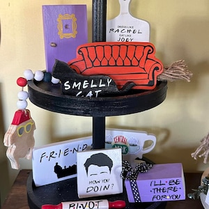 May include: A two-tiered black tray with wooden decorations themed after the TV show Friends. Includes a purple door, an orange couch, a black cat that says "Smelly Cat", and a white cutting board that says "Dress like Rachel, eat like Joey".