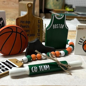 May include: A collection of wooden decorations with a basketball theme. The decorations include a basketball jersey, a basketball, a wooden sign that says "Eat Sleep Play", a wooden sign that says "Boston Celtics", a wooden sign that says "Go Team", and a wooden sign that says "I Love Basketball".