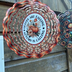 Op de afbeelding: Een decoratieve metalen windspinner met een herfstthema. Het midden heeft een pompoen- en bloemenontwerp met de tekst "Leaves are Falling, Autumn is Calling". De buitenste lagen hebben een golvend, gelaagd ontwerp in tinten oranje en bruin.