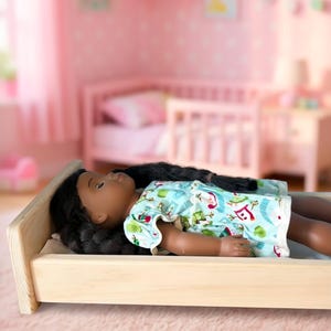 May include: A doll with dark skin and long black hair lying in a light-colored wooden bed. The doll wears a light blue short-sleeved shirt with a colorful pattern of small trees and red accents. The text "It's Bedtime !!!" is visible below the bed.