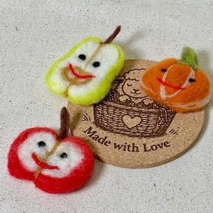 May include: Handmade felt fruit and vegetable decorations. Includes a red apple, a green pear, and an orange pumpkin, each with smiling faces. The decorations are on a cork coaster with the words "Made with Love".