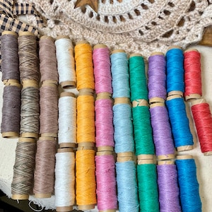 May include: A collection of thirty-one spools of waxed thread arranged in rows. The thread colors include shades of brown, white, yellow, pink, light blue, green, purple, bright blue, and red, all wound onto small cardboard cylinders.
