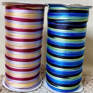 May include: Two cylindrical spools of satin cord. The left spool features alternating horizontal stripes of magenta, pale yellow, and white. The right spool features alternating horizontal stripes of dark blue, bright blue, and lime green.