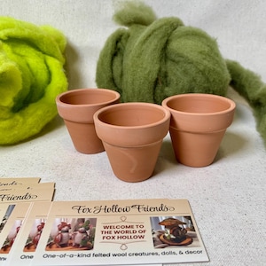 May include: Three small, empty terracotta pots are arranged in a row. Behind them are two large clumps of green and yellow wool roving. Several cards with the text "Fox Hollow Friends" and "Welcome to the World of Fox Hollow" are in the foreground.