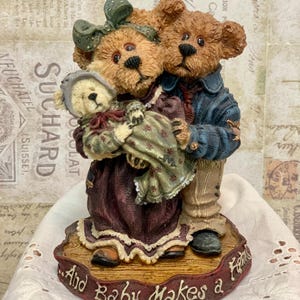 Pode incluir: Uma figura de uma família de ursos. A ursa mãe está segurando um ursinho em seus braços. O urso pai está de pé atrás deles. A base da figura é de madeira e tem o texto "And Baby Makes a Family" inscrito nela.