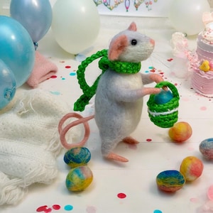 May include: A felted gray mouse wearing a green scarf, holding a green basket with a blue egg. The mouse has pink ears and a pink tail. Colorful decorative eggs and balloons are in the background.