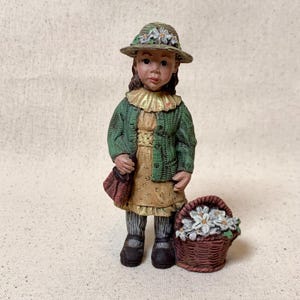 May include: A figurine of a young girl wearing a green cardigan, a floral hat, and a yellow dress. She carries a red purse and stands next to a basket filled with white flowers. The figurine has a vintage, handcrafted appearance.