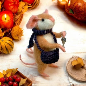 May include: A handmade felt mouse figure with a tan body, pink ears, and a white chest. It wears a navy blue scarf and bag, holding a silver charm. The scene includes pumpkins and candles, suggesting an autumn theme.