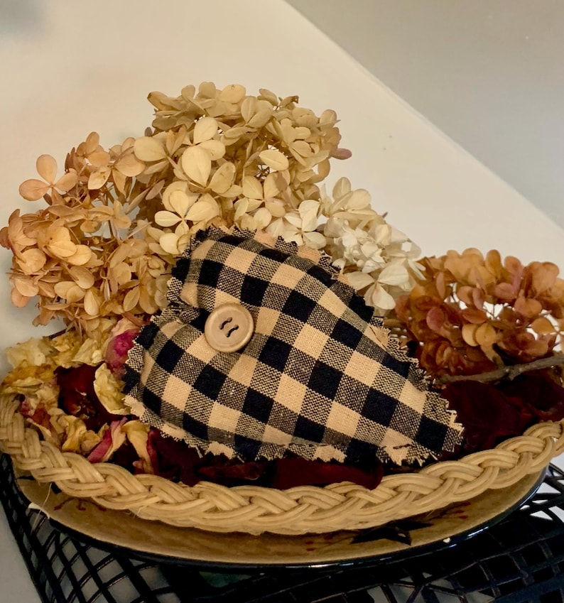May include: A black and tan checkered heart-shaped pillow with a wooden button in the center, sitting in a woven basket filled with dried flowers.