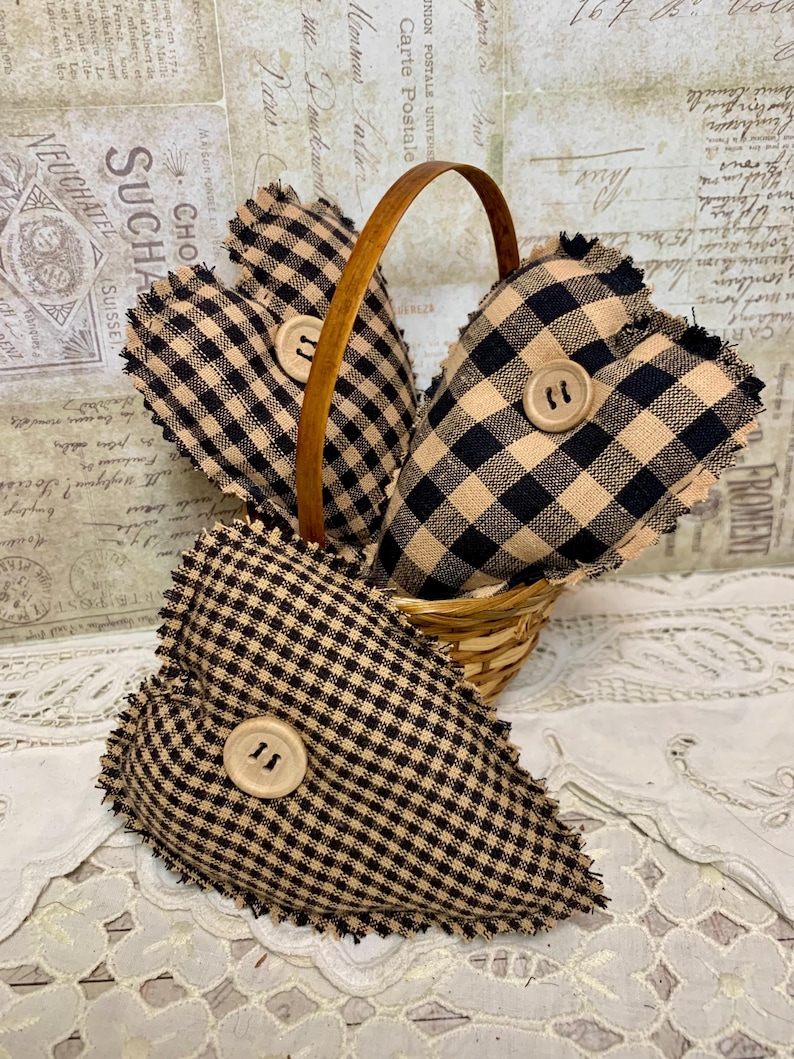 May include: Three black and tan gingham heart-shaped pillows with frayed edges and wooden buttons in a woven basket.