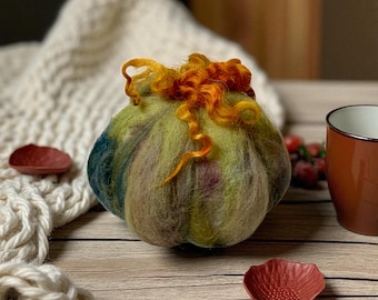 May include: A handmade, felted pumpkin with green, blue, and purple hues, topped with orange wool curls. The pumpkin sits on a wooden surface, accompanied by a small red dish and a brown mug.