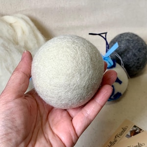 May include: A large, off-white felt ball is held in a hand, with other felt balls in the background. The felt ball is round and has a textured surface. Other felt balls are gray and white with blue accents. The theme is home decor.