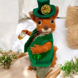 May include: A felt squirrel wearing a green hat with a gold shamrock, a green waistcoat with a shamrock pin, and green and grey trousers. The squirrel is holding a glass of beer.
