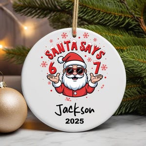 May include: A white ceramic Christmas ornament with the text "SANTA SAYS" and a cartoon Santa Claus wearing sunglasses. The ornament also includes the name "Jackson" and the year "2025". A gold-colored ornament is in the background.