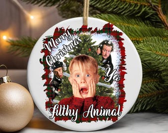 May include: A white ceramic Christmas ornament with a festive design. The ornament features a central image of a child with a surprised expression, surrounded by text that reads "Merry Christmas Ya Filthy Animal". A gold-colored ornament is in the foreground.