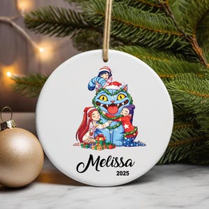 May include: A white ceramic Christmas ornament featuring a cartoon illustration of a blue cat wearing a Santa hat, surrounded by children. The ornament has the name "Melissa" and the year "2025" printed on it. A gold-colored ornament is next to it.