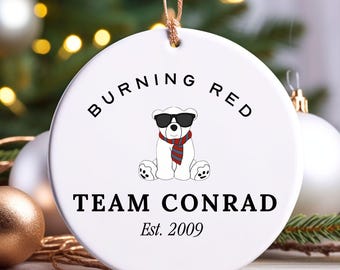 May include: White ceramic Christmas ornament with the text "BURNING RED" and "TEAM CONRAD Est. 2009". The ornament features a cartoon polar bear wearing sunglasses and a scarf. The ornament is hanging in front of a Christmas tree.