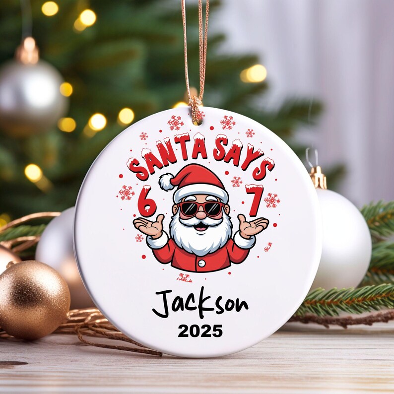 May include: A white, round Christmas ornament with a cartoon Santa Claus wearing sunglasses. The text "SANTA SAYS 6 7" is above the Santa graphic. Below the graphic is the name "Jackson" and the year "2025". The ornament hangs from a gold string.