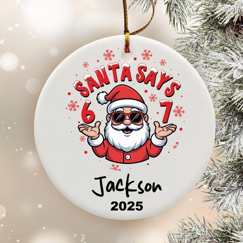 May include: A white Christmas ornament with a cartoon Santa Claus wearing sunglasses. The text "SANTA SAYS 6 7" is above the Santa graphic. Below the graphic is the name "Jackson" and the year "2025". A gold string is attached.