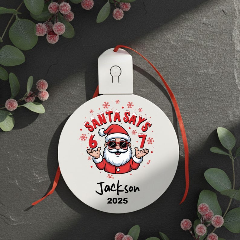 May include: White Christmas ornament with a red ribbon. The ornament features a cartoon Santa Claus wearing sunglasses and the text "SANTA SAYS 6 7". Below the Santa graphic is the name "Jackson" and the year "2025".