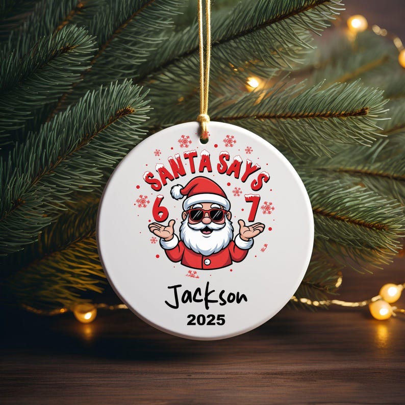 May include: A white Christmas ornament with a cartoon Santa Claus wearing sunglasses and a red suit. The ornament reads "SANTA SAYS 6-7" and "Jackson 2025". The ornament is hanging from a gold string, with a Christmas tree in the background.
